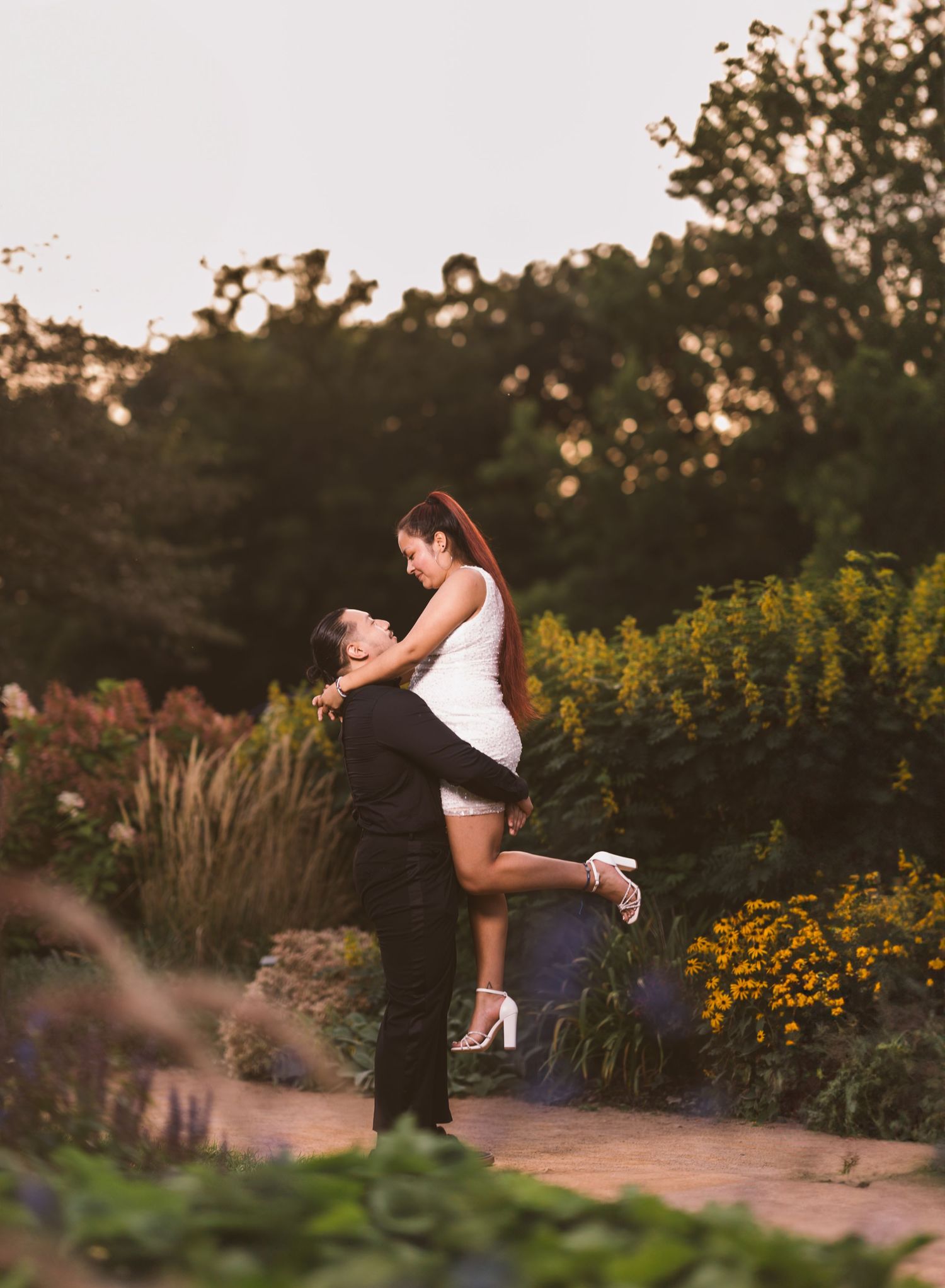 A couple embraces in a romantic dance pose surrounded by summer garden flowers at sunset.