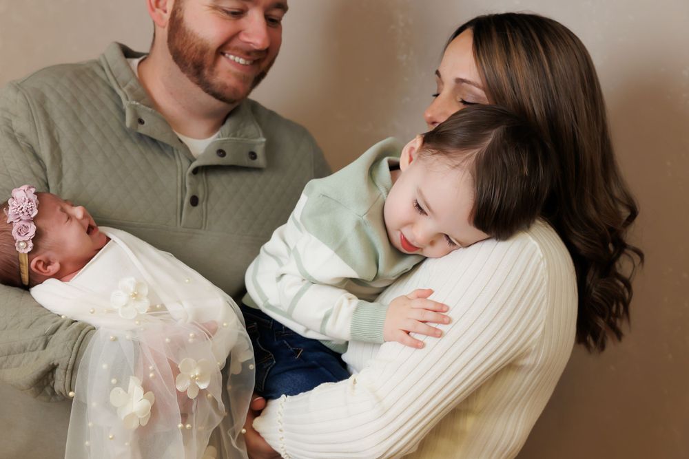 A tender family moment shows parents with their newborn baby and young child in warm, casual indoor setting.