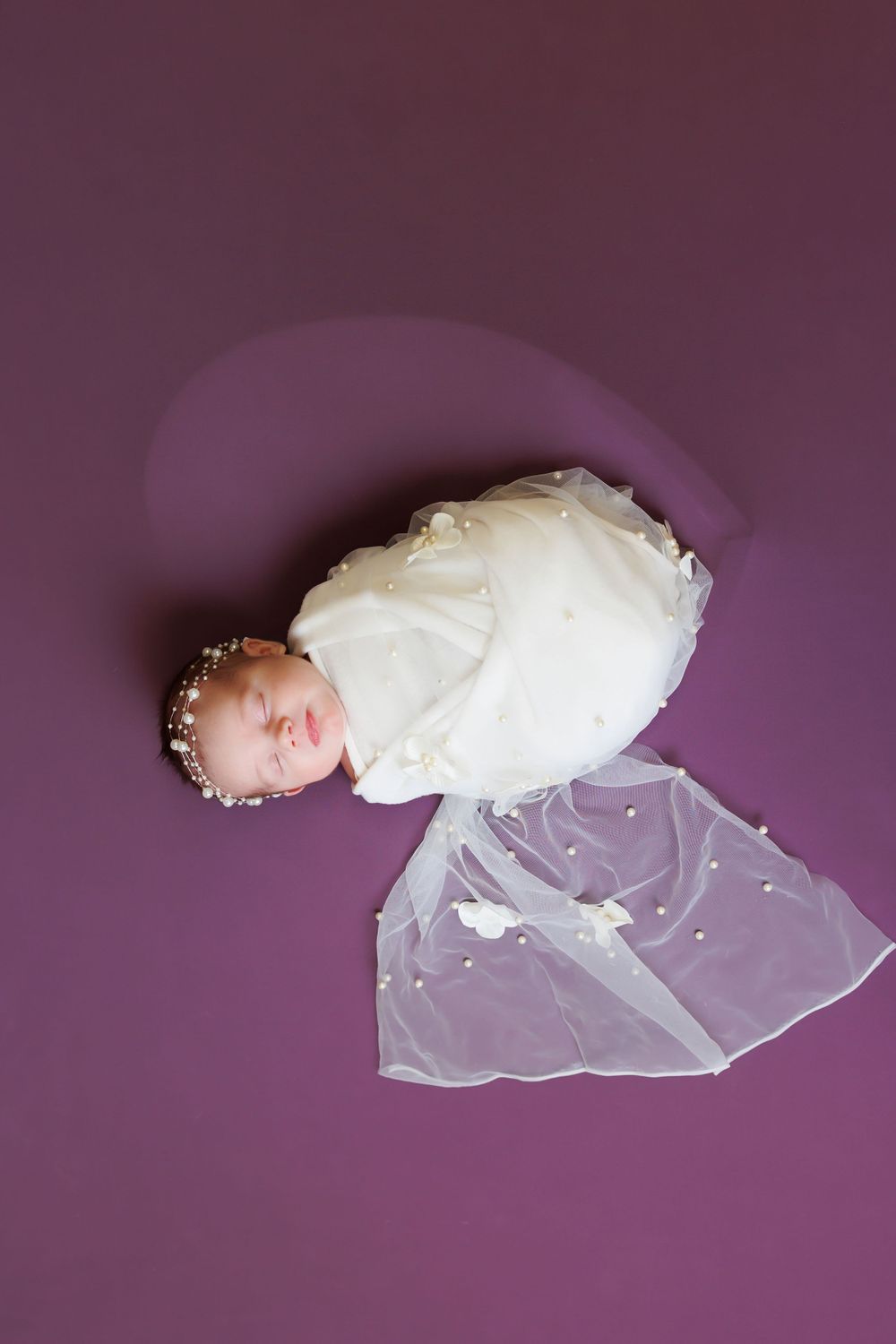 Newborn in white dress and headband lying on purple background with lace veil.