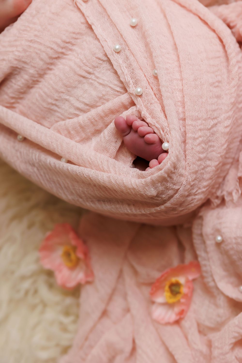 Tiny baby foot peeking out of pink textured blanket with pearl buttons and floral accents.