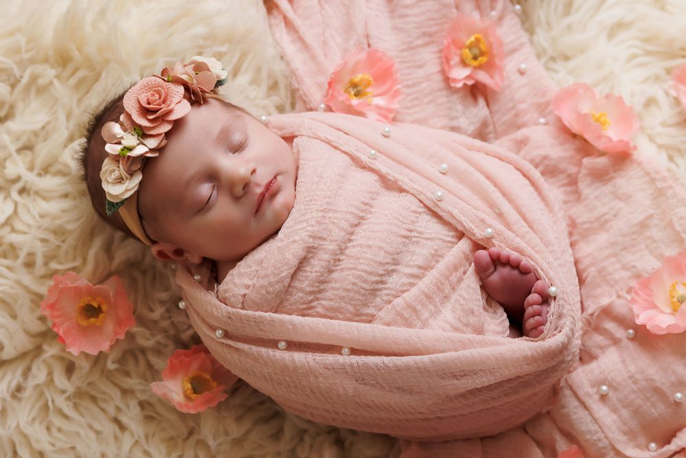 Peaceful newborn wrapped in pink swaddle surrounded by coral flowers on cream fur.