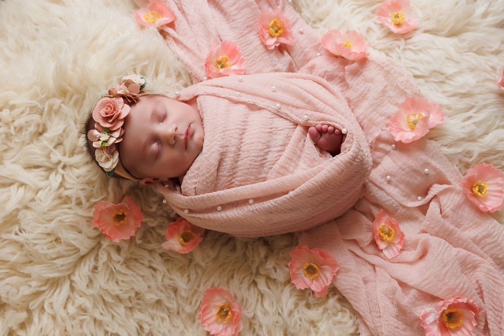 A sleeping newborn in pink wrap surrounded by coral flowers on cream textured backdrop.