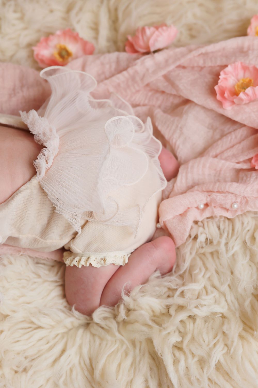 Delicate detail of baby's feet in white ruffled dress on cream fur with pink flowers.