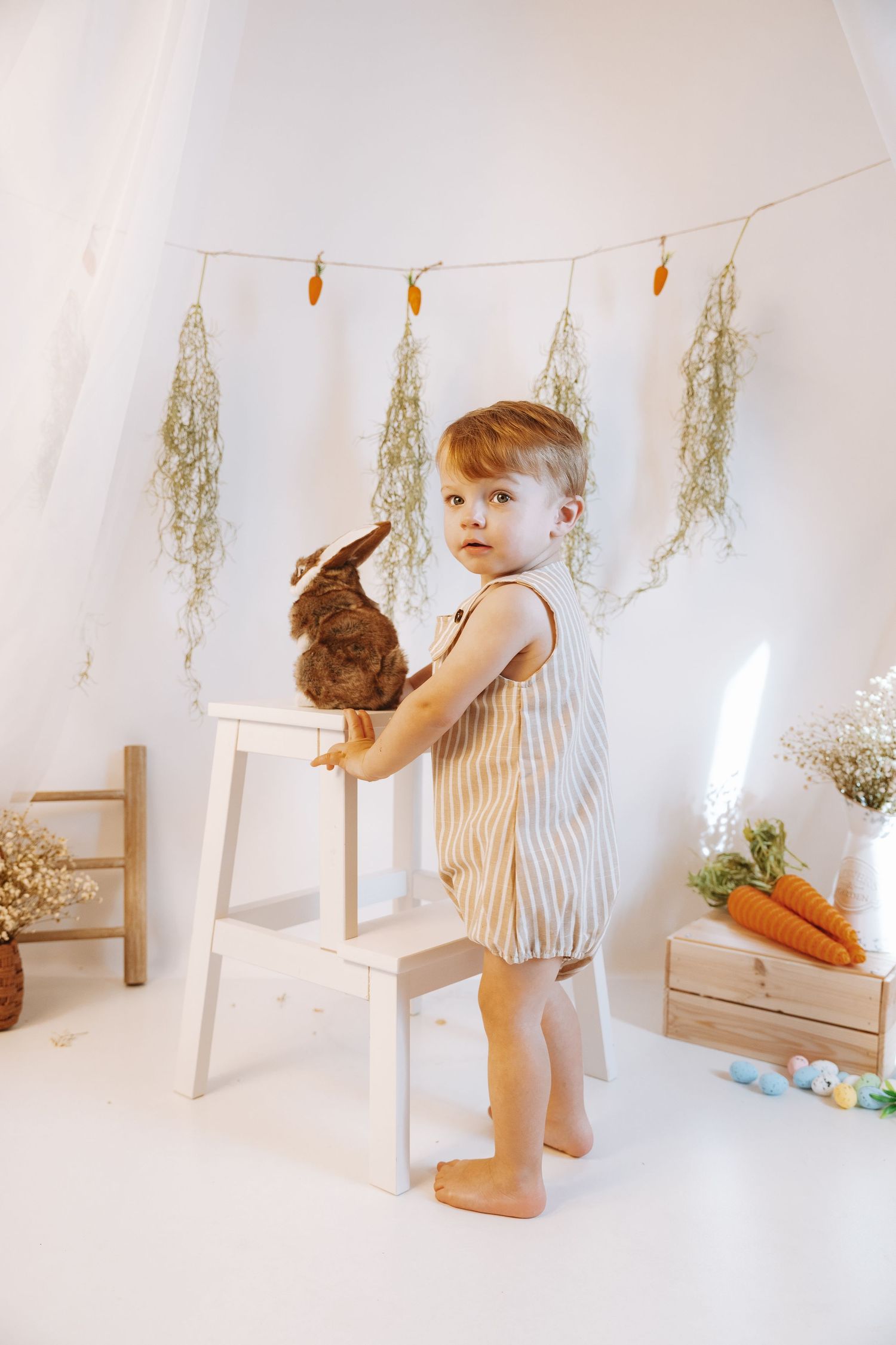 A small child in a striped dress holds a plush bunny rabbit beside a white chair with dried flowers hanging above.