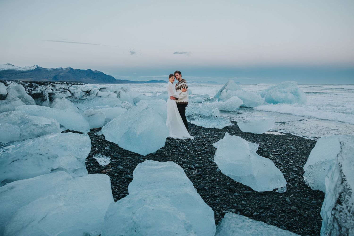 A couple embraces among crystal blue ice formations on a black sand beach near Jokulsarlon Glacier Lagoon in Iceland.