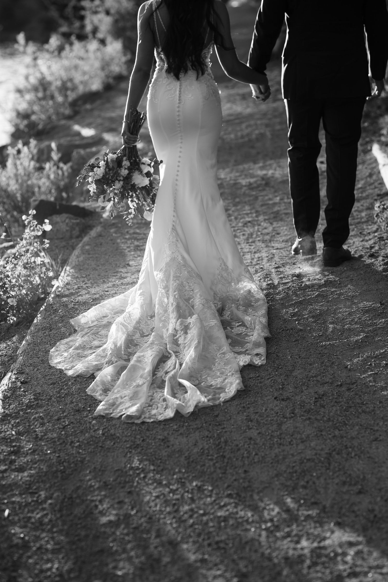 Black and white wedding photo showing flowing lace wedding dress train as couple walks hand in hand on rustic path.