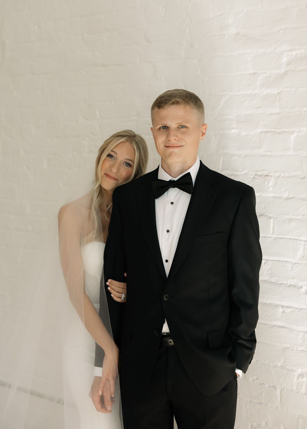 A couple in formal wedding attire poses against a white brick wall in a series of romantic shots.