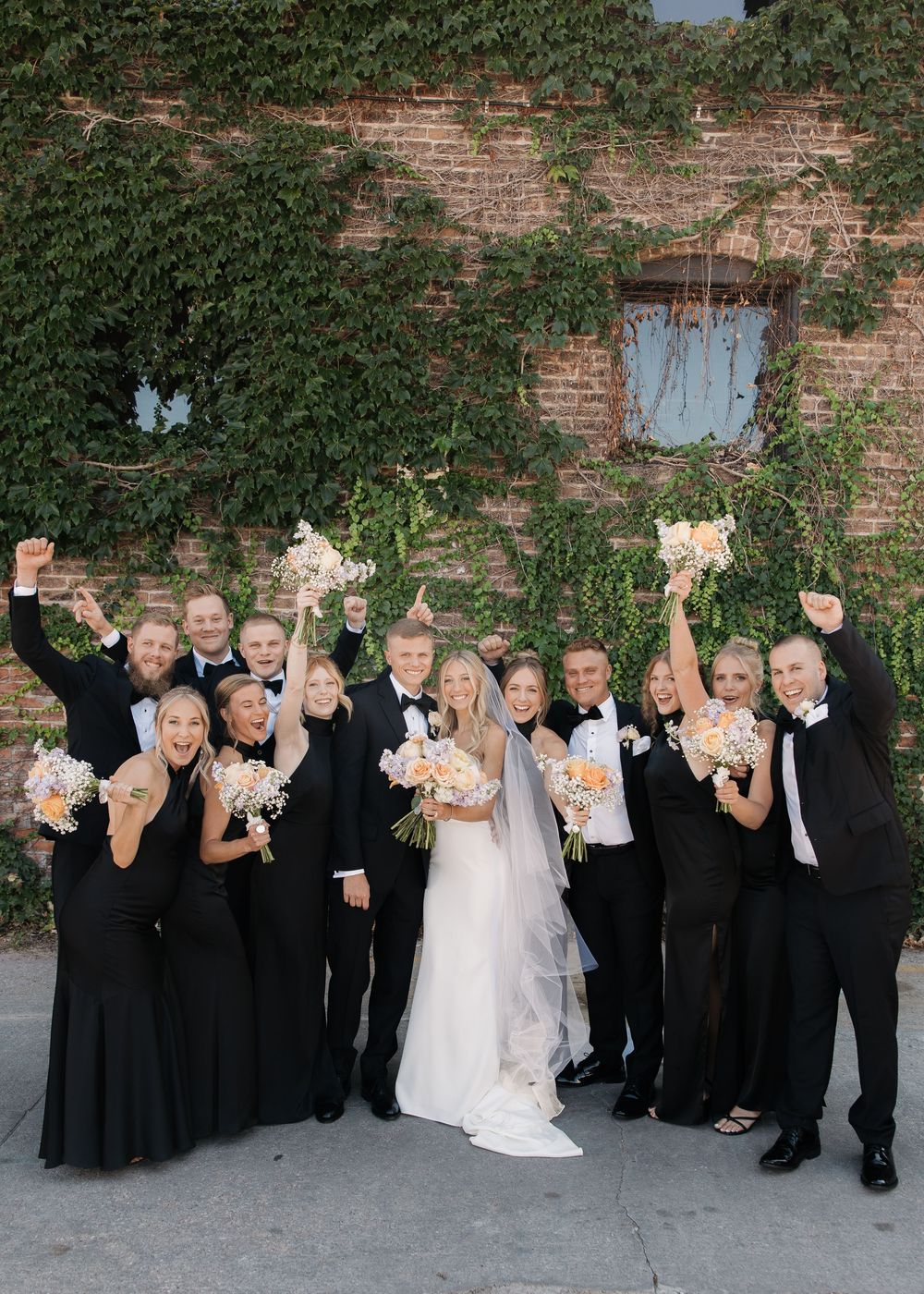 Wedding party in black attire holding up paper bags in a fun group photo against a brick wall with ivy.
