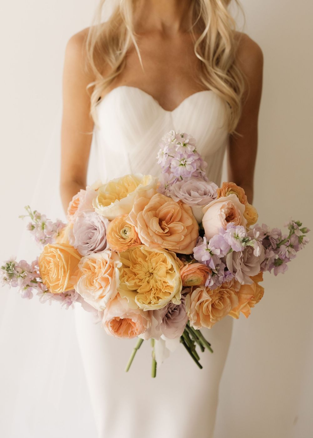 A delicate floral arrangement featuring peach garden roses, lavender blooms, and soft greenery against a white background.