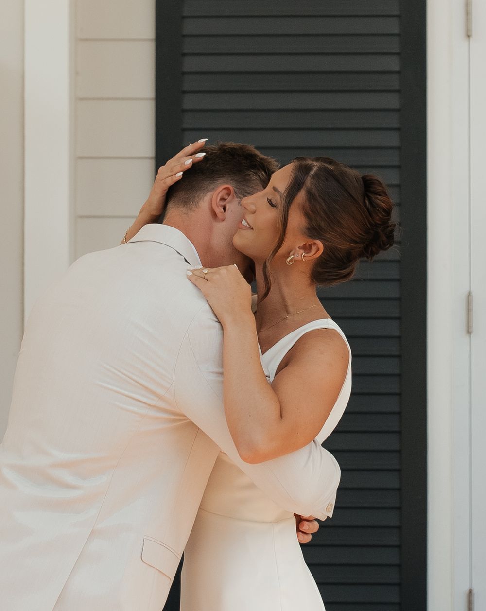 A couple in white attire share three romantic kisses against a backdrop of dark shutters.