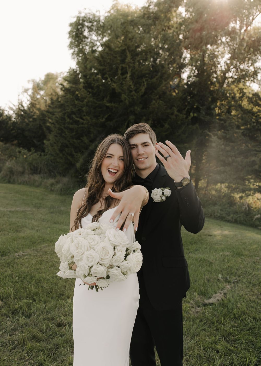 A romantic couple poses together outdoors during sunset in a grassy field with white roses.