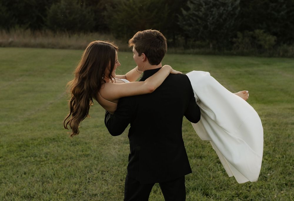 A couple shares a romantic moment in a grassy field as the person in black lifts their partner wearing flowing white.