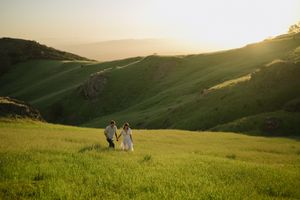 Two people walking together across a lush green hillside at golden hour with rolling mountains in the background.