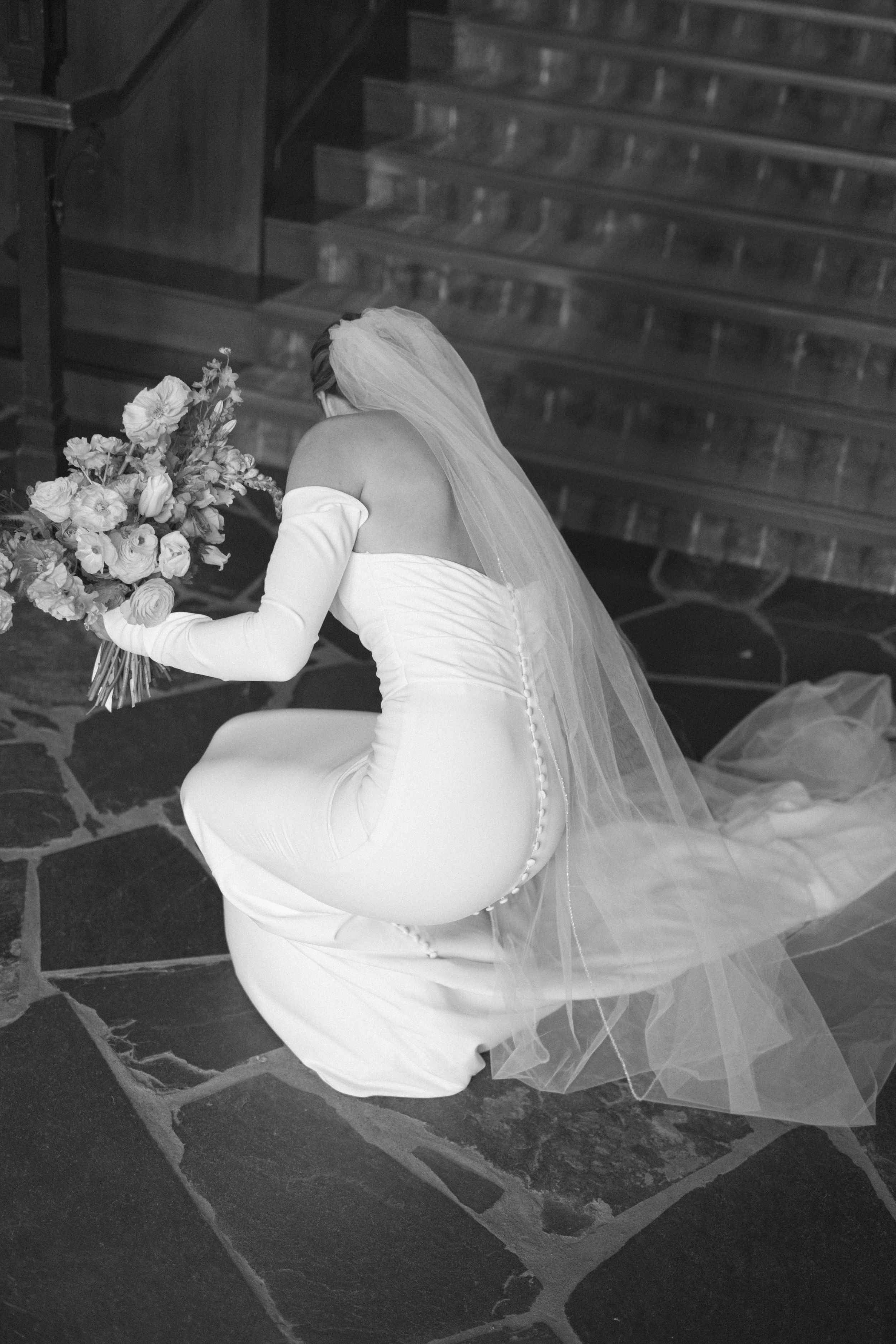 A bride in a white satin dress with a long veil kneels on stone steps while holding a floral bouquet in black and white.