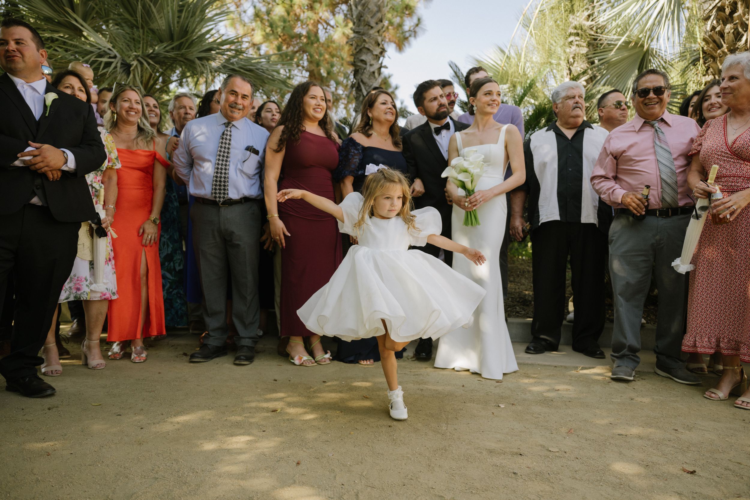 Wedding guests dance and celebrate on an outdoor dance floor surrounded by palm trees during a daytime celebration.