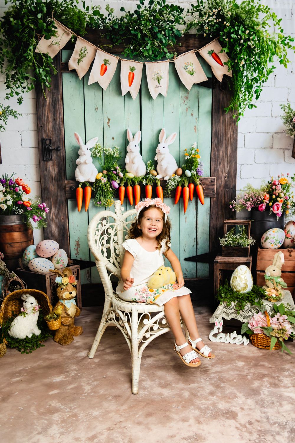 Spring photo backdrop with white wicker chair, Easter bunnies, carrots, and decorative banners against mint green door.