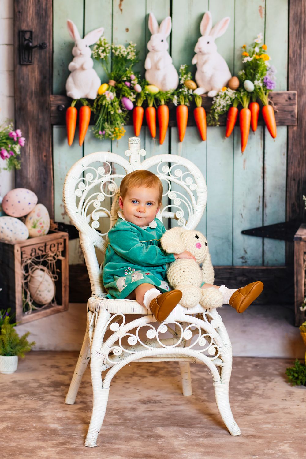 Easter photo setup with white bunnies and carrots on rustic wall above ornate white wicker chair with plush decorations.