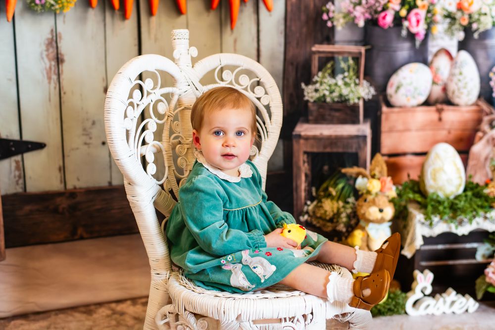 A small figure in teal green sits in an ornate white chair surrounded by Easter decor and spring flowers.