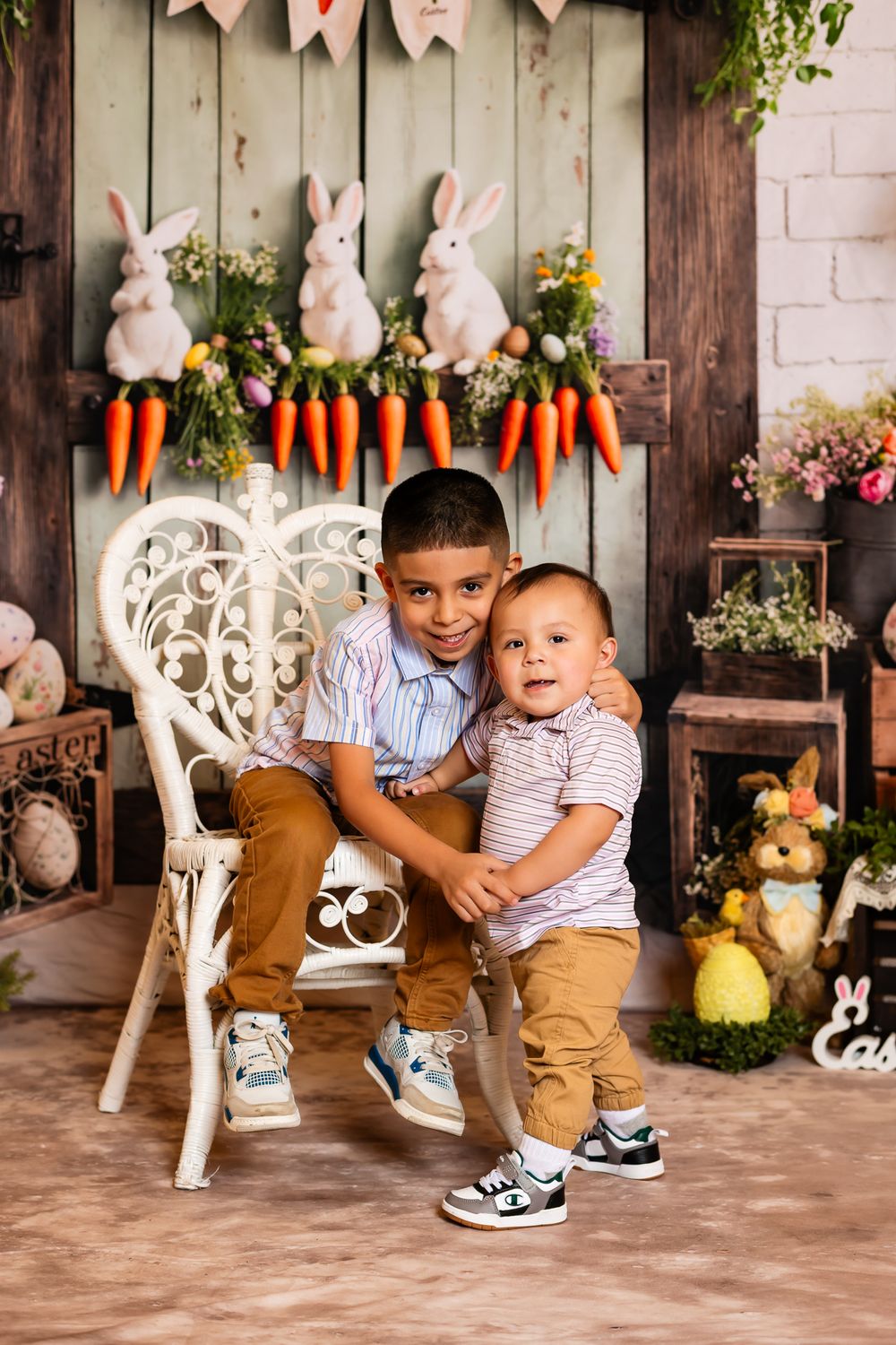 Easter photo setup with white bunnies, decorative carrots, and a white wicker chair against a rustic wooden backdrop.