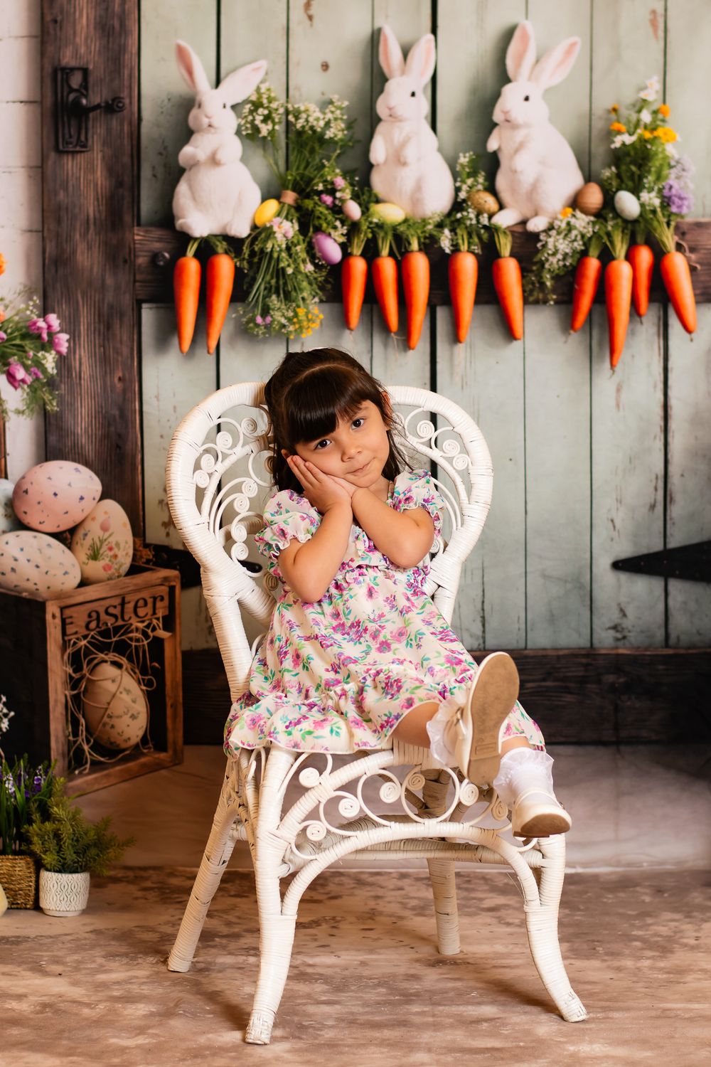 Spring Easter photo setup with white wicker chair, decorative bunnies and carrots on rustic wooden wall backdrop.
