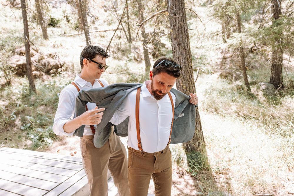 Two people in dress attire help adjust each other's outfits while standing on a wooden deck surrounded by pine trees.