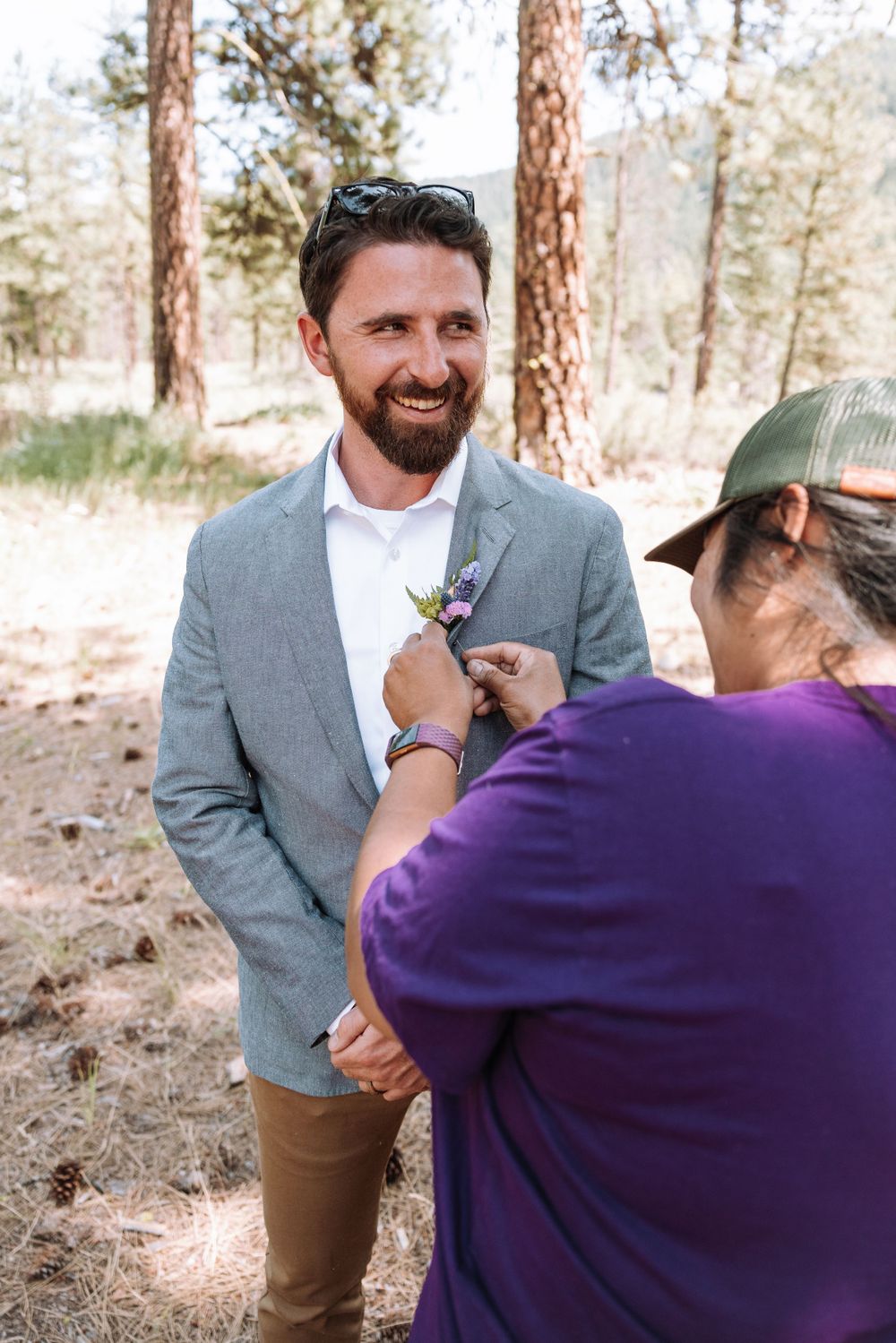 A wedding preparation moment as someone pins a boutonniere onto a grey suit jacket in a pine forest setting.