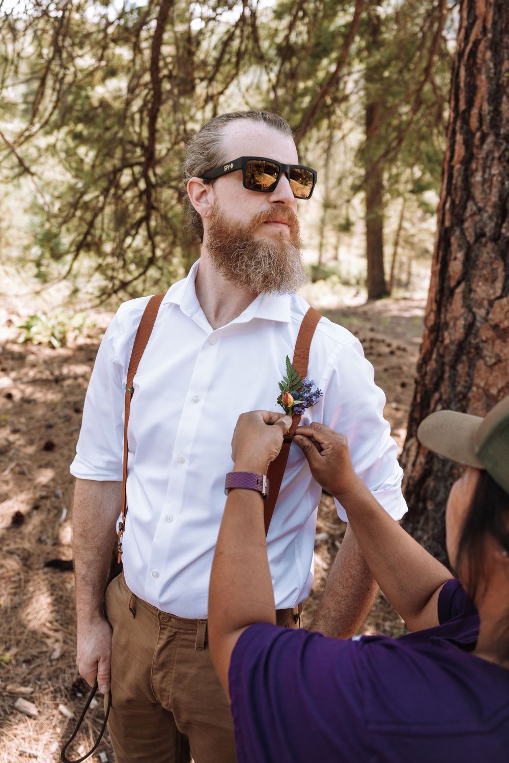 A bearded man in suspenders and sunglasses gets boutonniere pinned to his white shirt in a forest setting.
