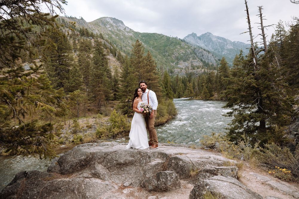 A romantic wedding portrait on rocky outcrop overlooking mountain river and forest landscape.