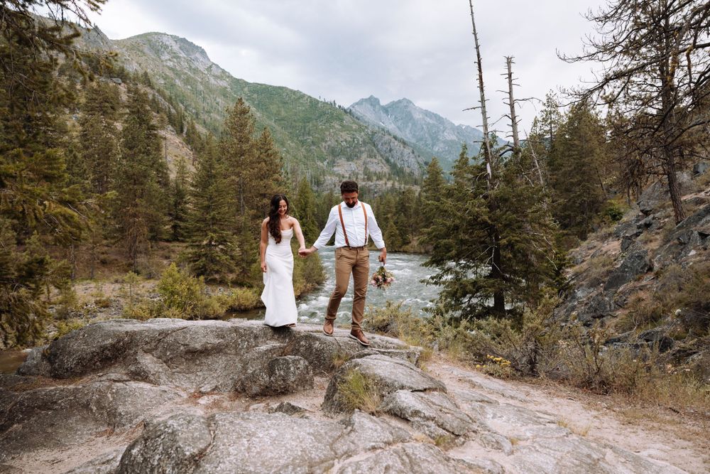 A couple stands on rocky terrain with dramatic mountain views in the background.