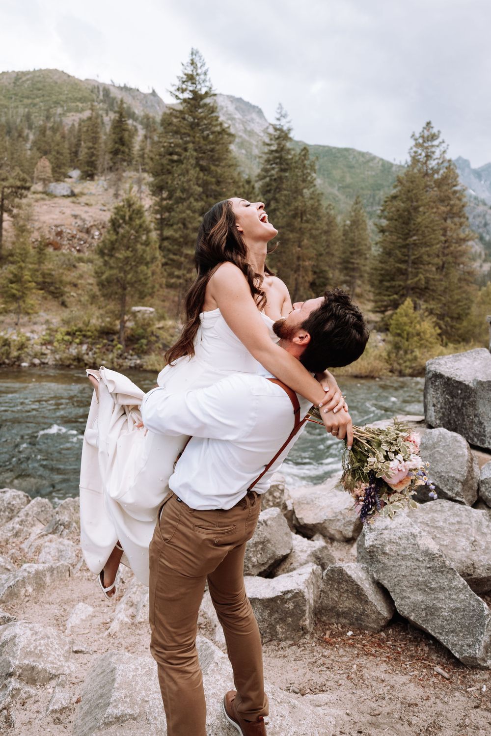 A playful wedding couple poses on rocks by a rushing mountain river.