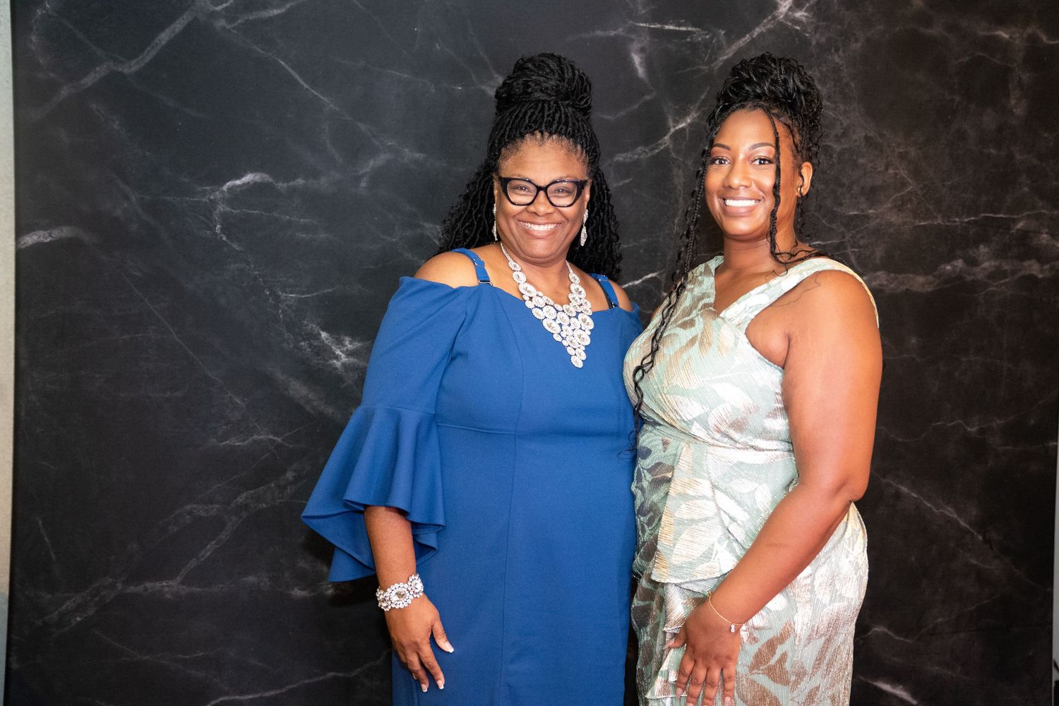 Two people in formal evening wear pose together against a dark marble background.