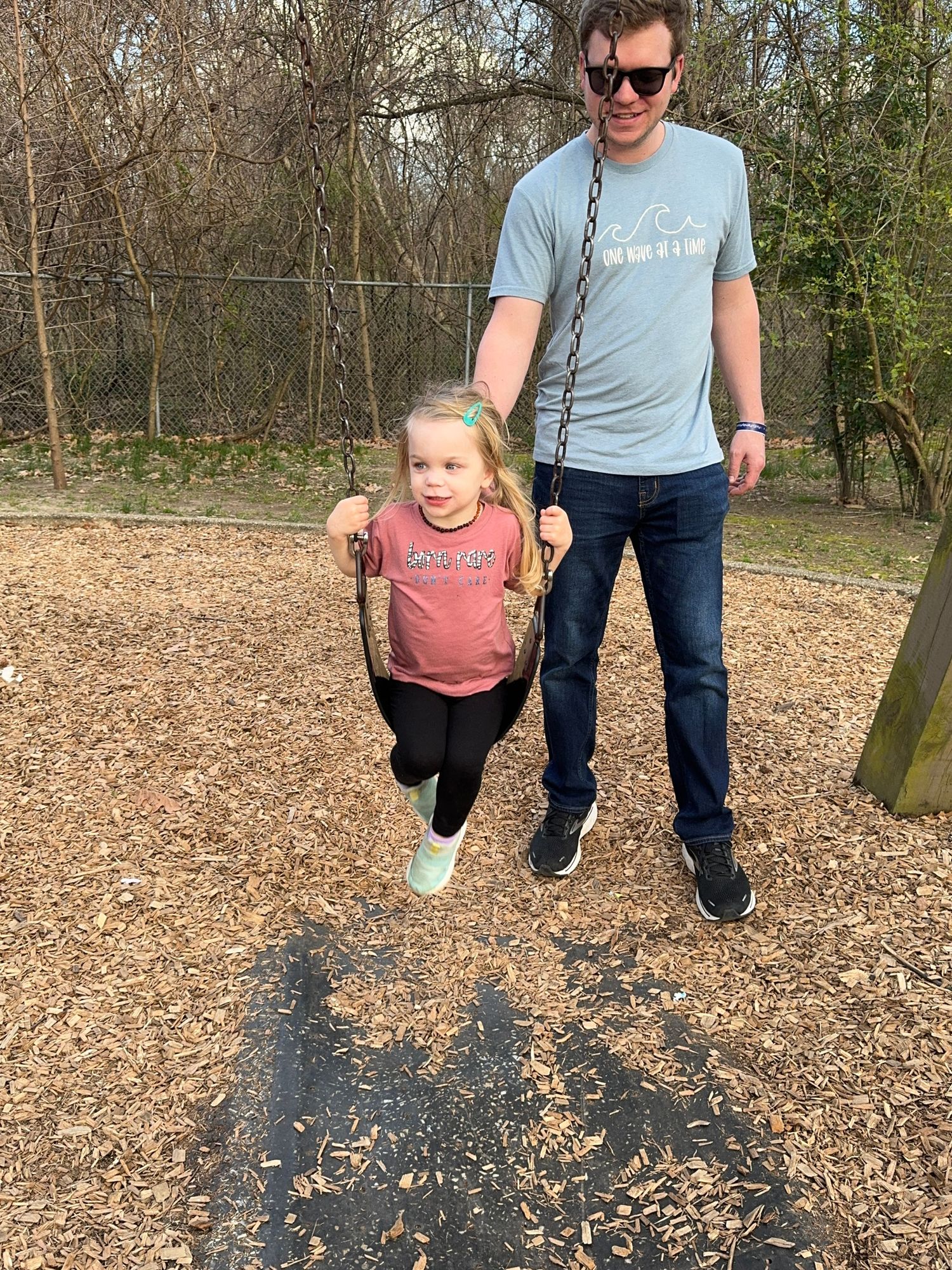 People enjoying a sunny day at a playground with mulch ground cover and trees in the background.