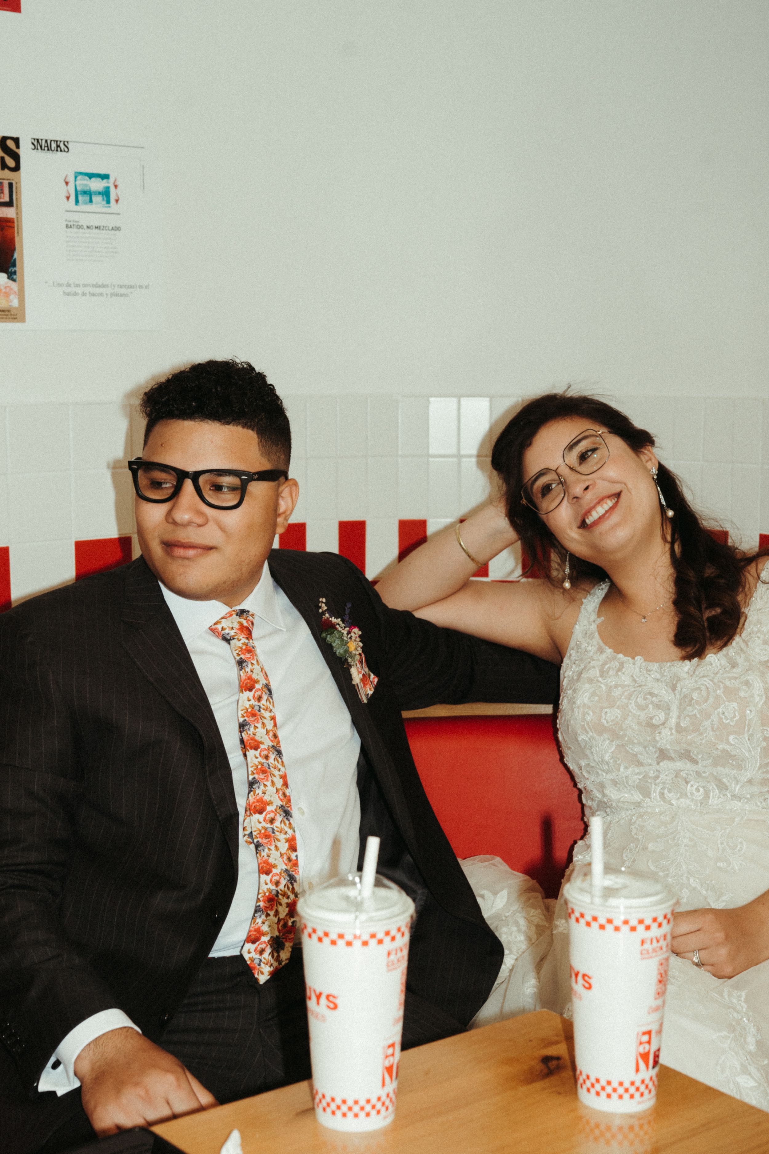 A couple in wedding attire sharing milkshakes at a casual restaurant with red decor.