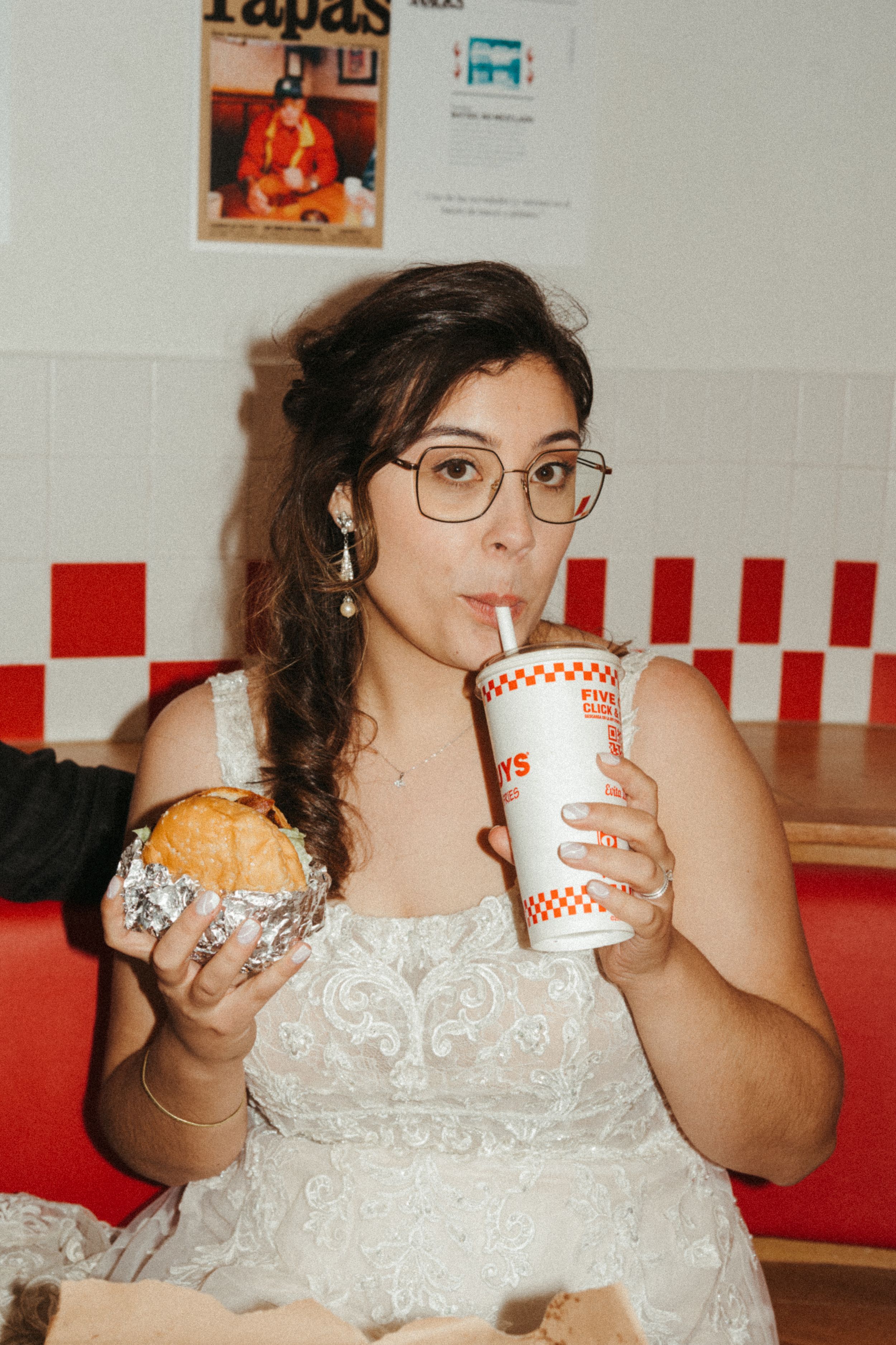 Someone in a white wedding dress enjoys fast food with a burger and drink at a casual restaurant with red and white decor.