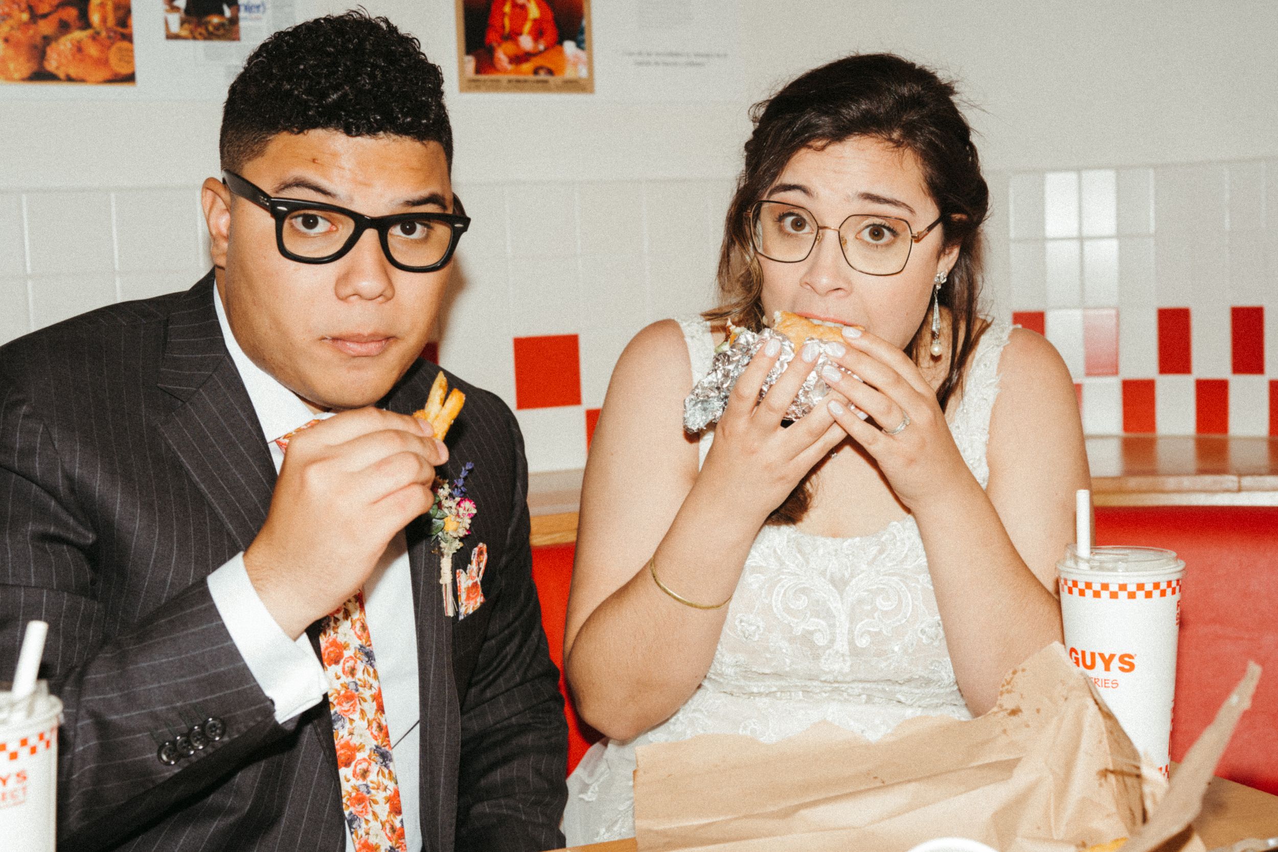 Newlyweds in matching glasses enjoy burgers together at a retro diner with red and white tile decor.