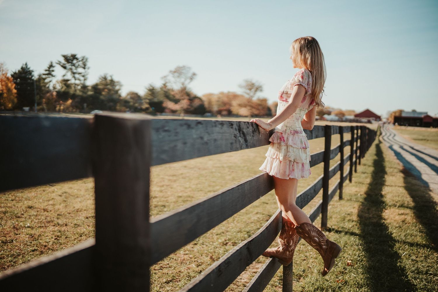 Girl in a floral dress leans against wooden fence at sunset while golden light casts long shadows on grassy field.