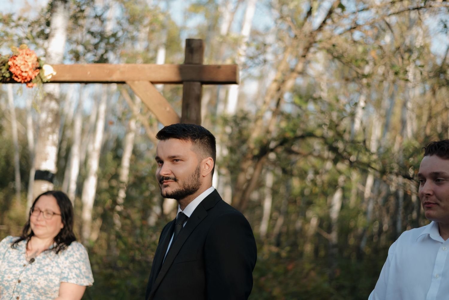 People gathered outdoors in front of a wooden cross during a wedding ceremony in a forest setting.