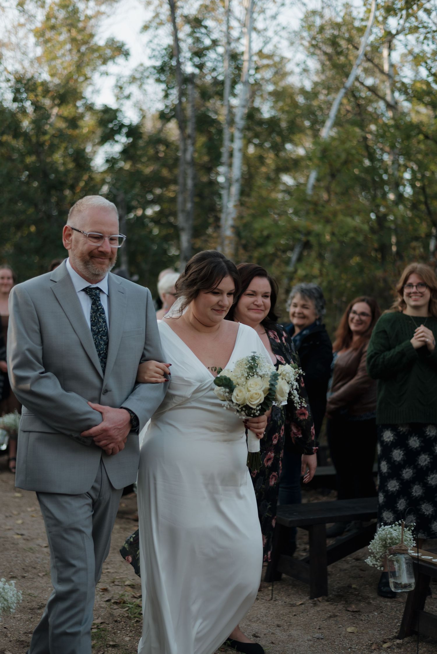 An outdoor wedding ceremony shows a couple walking down a dirt path while guests look on from the side among trees.