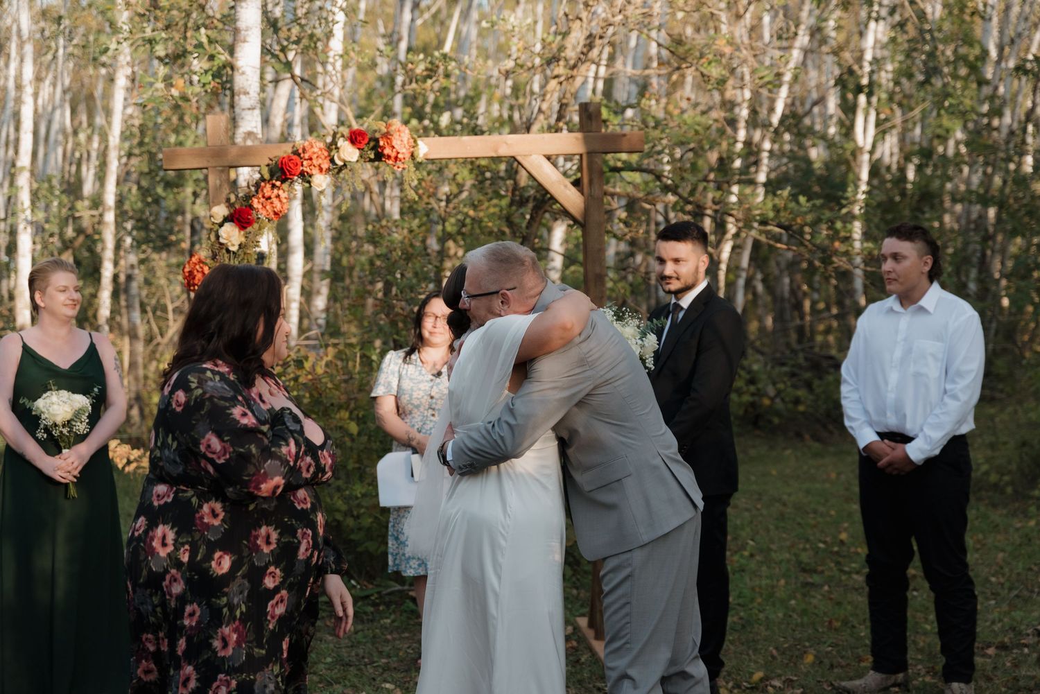 An intimate outdoor wedding ceremony takes place under a wooden arch decorated with autumn flowers and greenery.