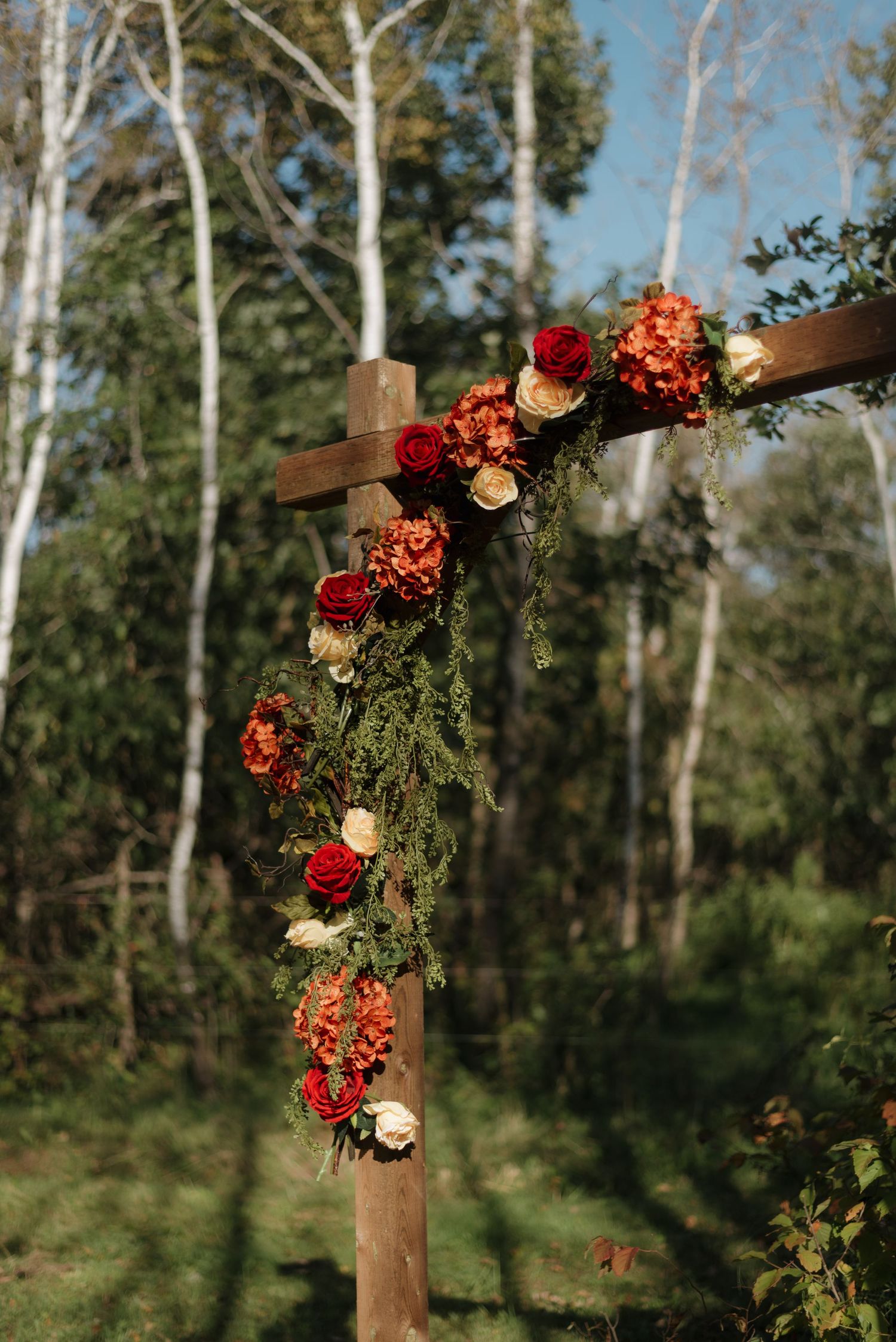 A wooden cross decorated with red roses and orange flowers against a backdrop of birch trees in a forest setting.