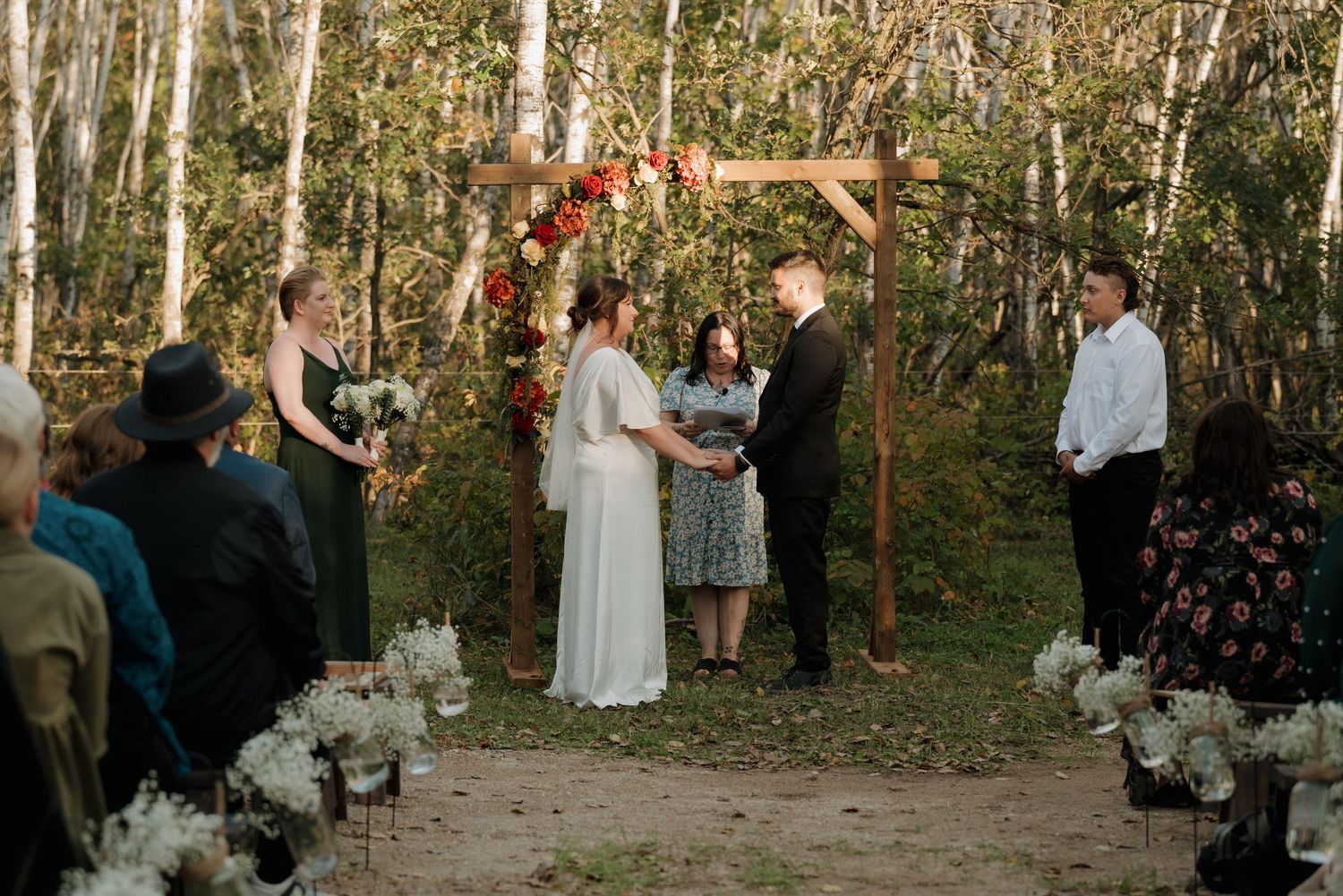 A rustic outdoor wedding ceremony takes place under a wooden arch adorned with autumn florals in a birch forest setting.