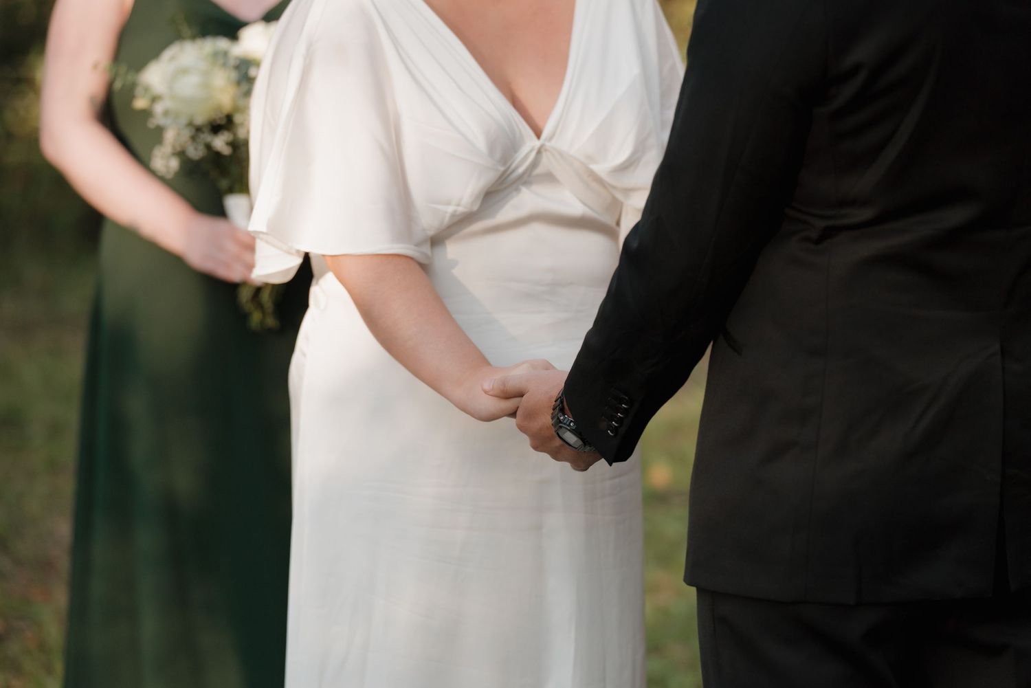 Close-up detail of couple holding hands during outdoor wedding ceremony with white dress and black suit.