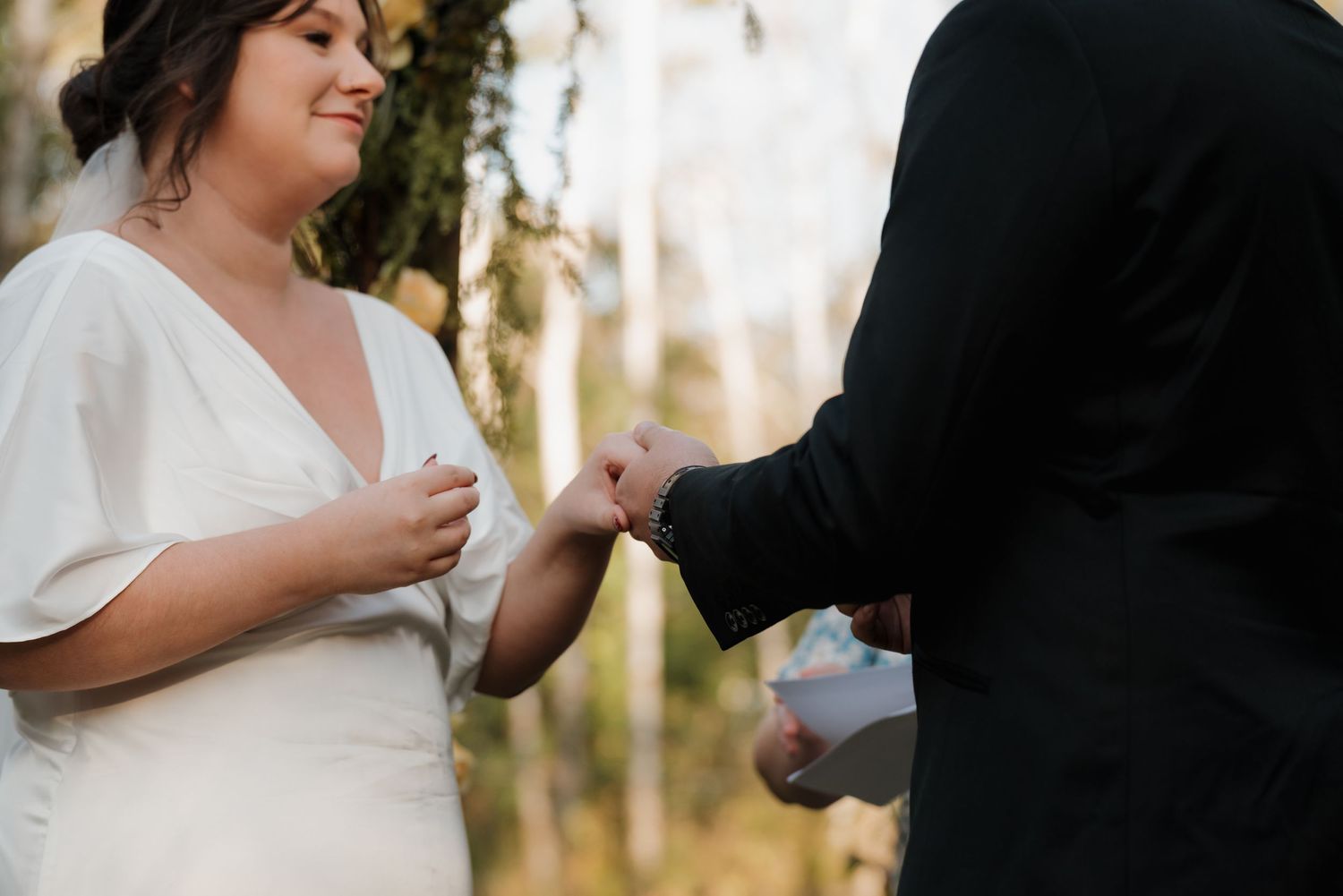 A romantic moment during a wedding ceremony shows hands exchanging rings in an outdoor woodland setting.
