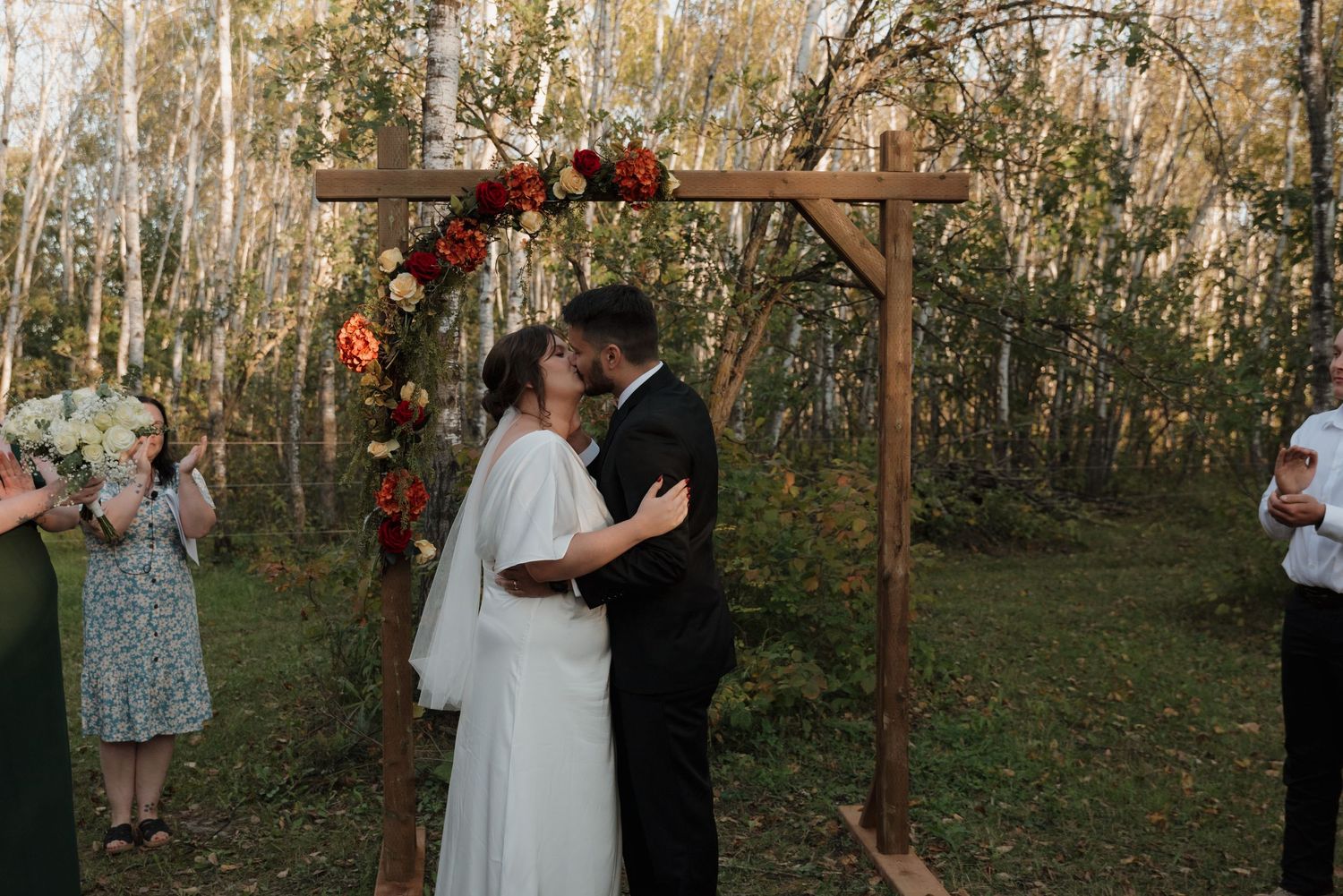 A couple shares their first kiss under a rustic wooden arch adorned with red and pink flowers at an outdoor autumn wedding.