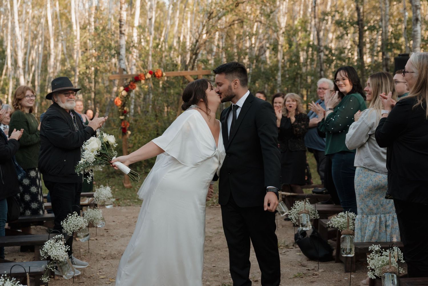 A couple celebrates during their outdoor forest wedding ceremony with birch trees and guests in the background.