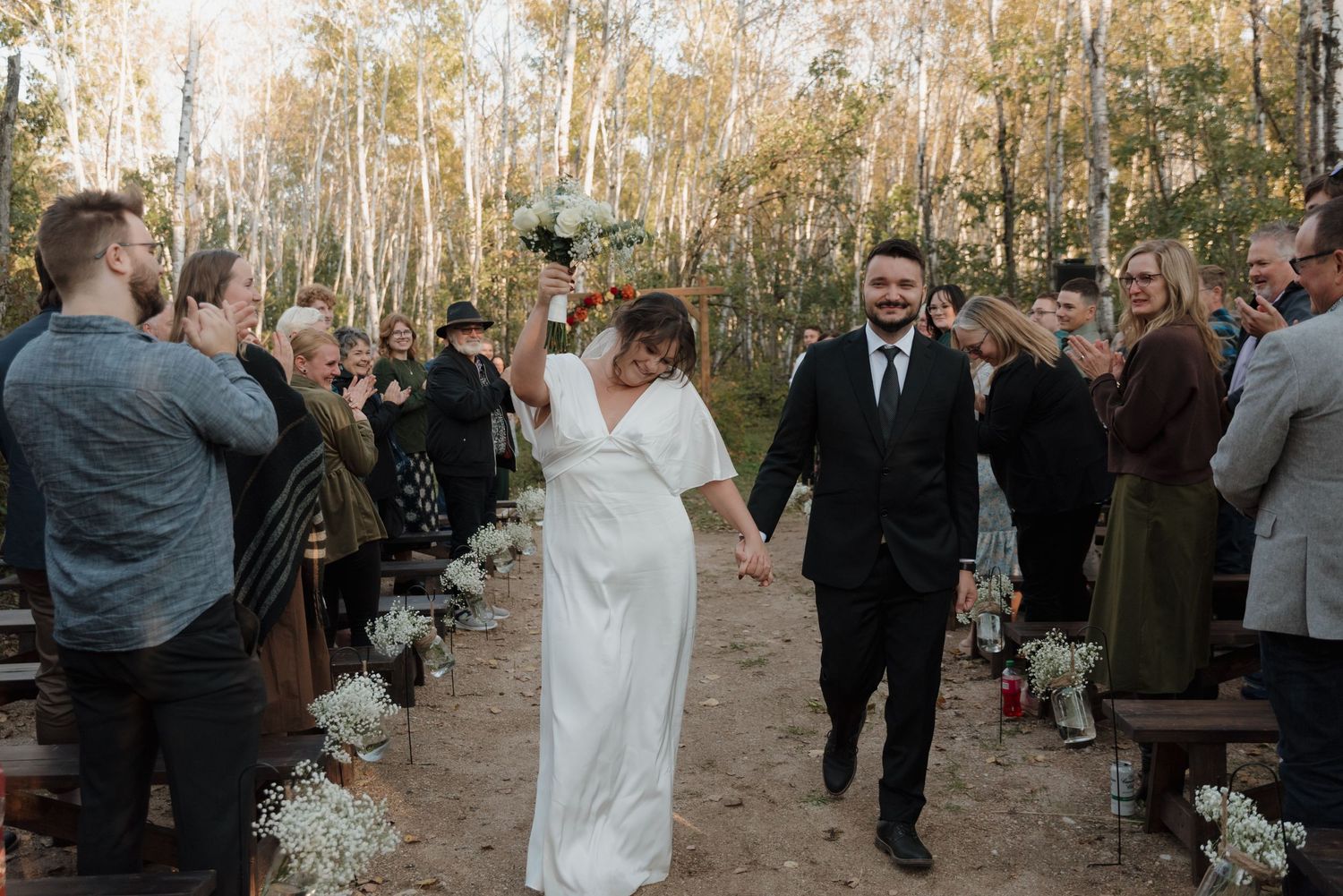 A joyous couple walks down an outdoor aisle lined with guests and white floral arrangements in a birch tree forest.