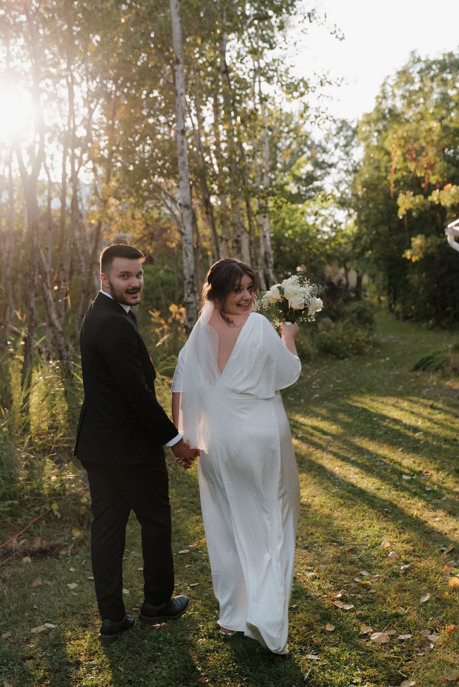 A wedding couple stands in golden sunlight filtering through autumn birch trees at sunset.