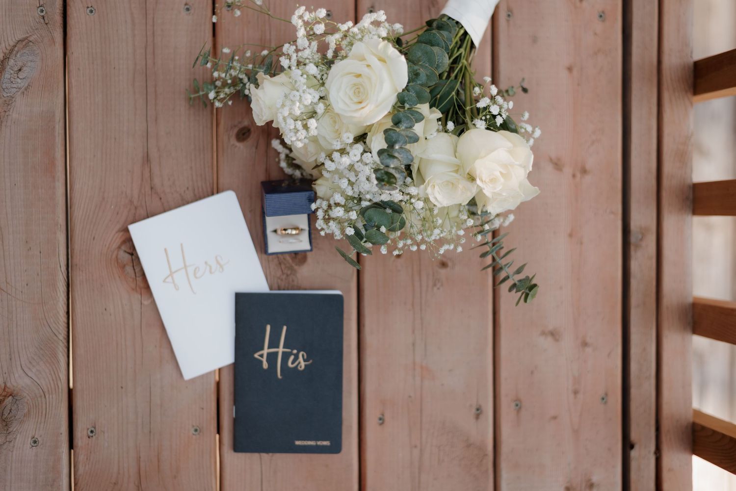 Wedding details featuring a navy blue vows book, white flowers, and invitations arranged on rustic wooden surface.