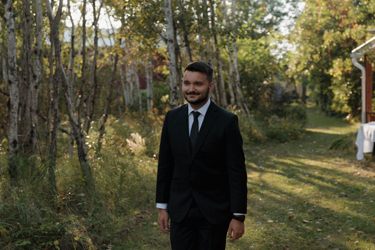 Someone in a black suit stands on a grass path surrounded by birch trees on a sunny autumn day.