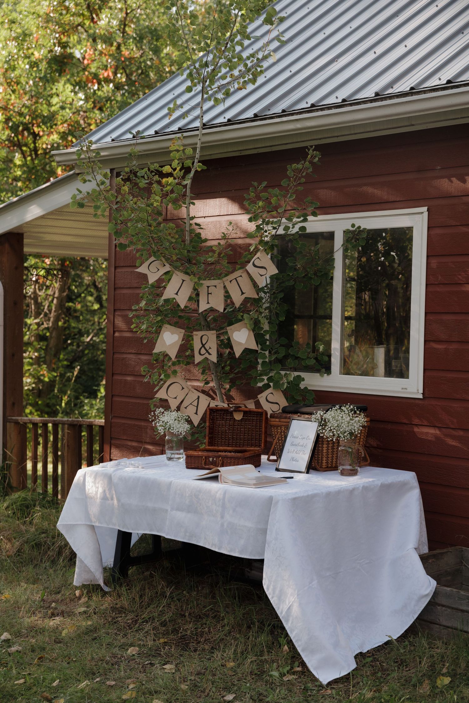 Rustic wedding reception table with white tablecloth and burlap banner decorations against red barn exterior.